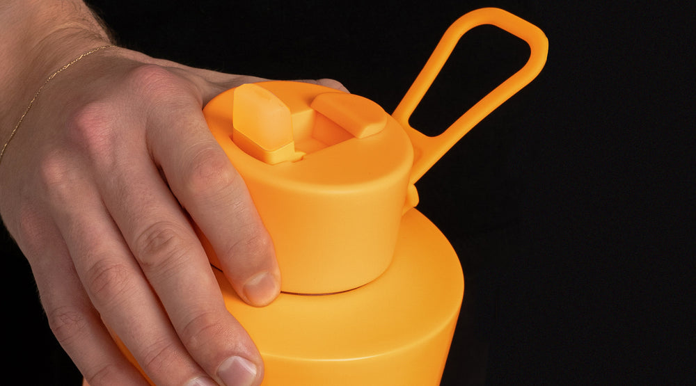 A close-up of a hand pressing the lid of a bright orange reusable water bottle against a solid black background.