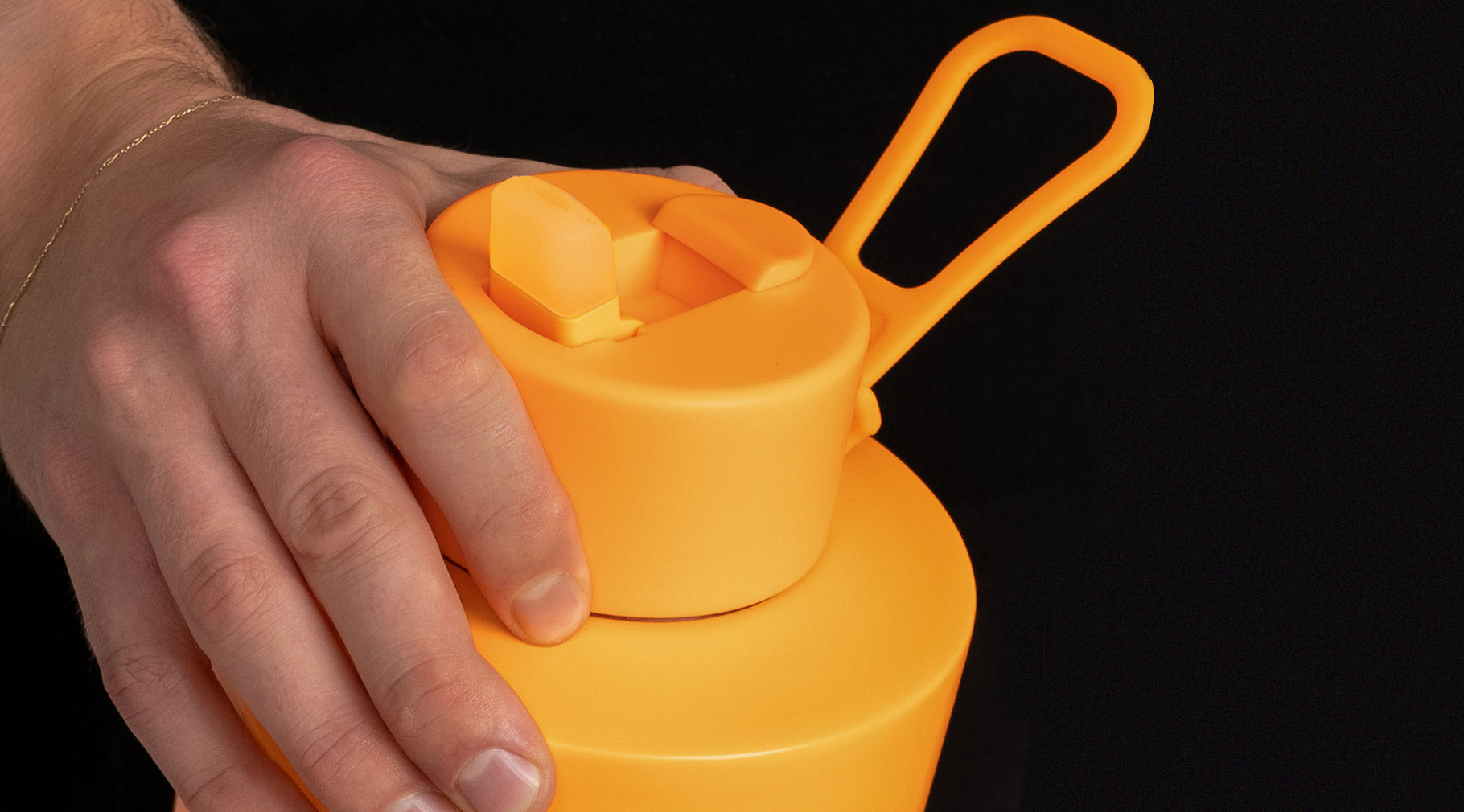 A close-up of a hand pressing the lid of a bright orange reusable water bottle against a solid black background.