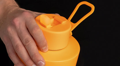 A close-up of a hand pressing the lid of a bright orange reusable water bottle against a solid black background.