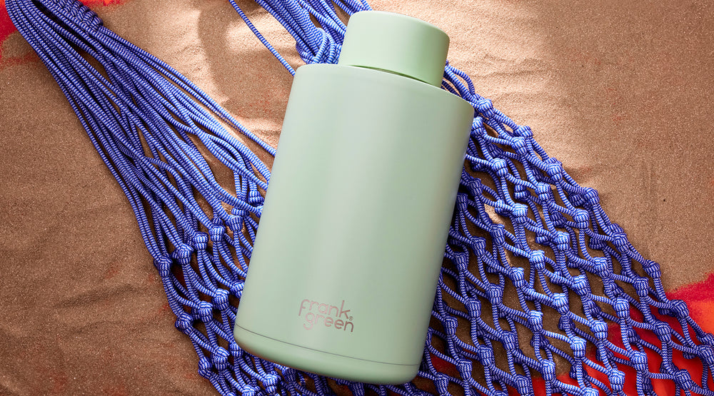 A mint green insulated bottle with a matching lid, featuring the 'frank' brand logo, resting on a blue woven hammock over a brown textured blanket.