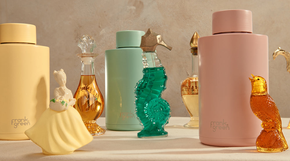 A collection of Frank Green glassware and bottles arranged on a tabletop against a textured beige wall. The set includes a large pale yellow bottle, a small yellow dress-shaped bottle, a mint green bottle with a gold seahorse topper, a translucent teal seahorse bottle, a large dusty pink bottle, and amber glass bottles with gold caps.