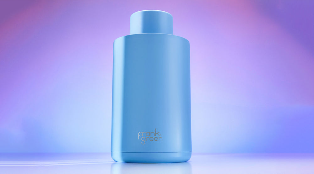 A light blue cylindrical bottle with a matching cap, featuring the 'crank green' logo near the bottom, set against a soft purple and blue gradient background.